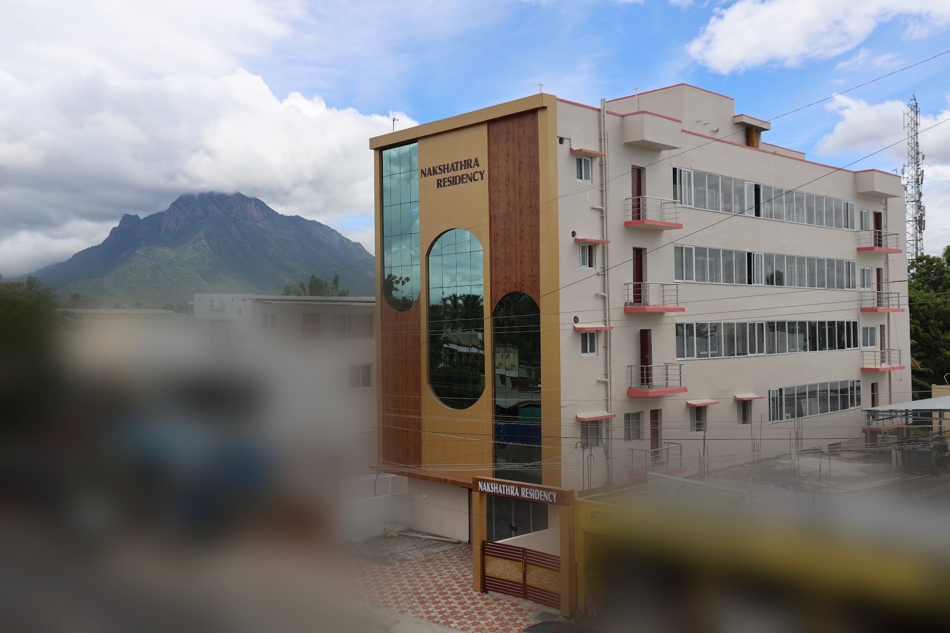 Nakshathra Residency, a five-storey building with a tall glazed entrance tower, photographed from the road with a rocky hill rising behind it.