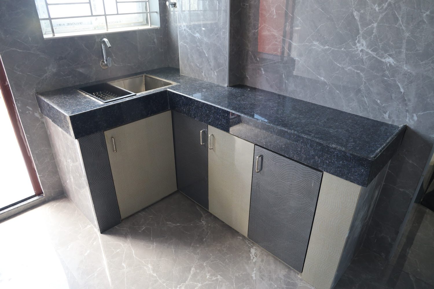 An L-shaped kitchen counter in black granite with a stainless sink and cupboards below, lit by a window.
